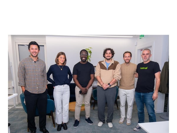 A group portrait featuring six people (five men and one woman) standing side-by-side and smiling in a modern, light-filled office or co-working space.