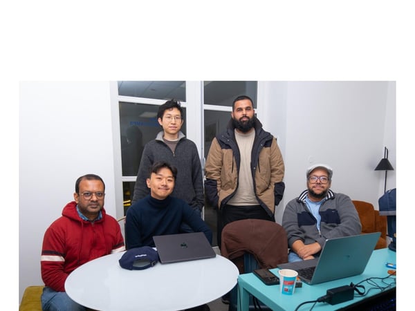 A group portrait of five men gathered indoors around two tables with laptops, likely in an office or co-working space.