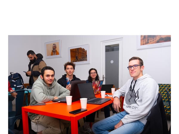 A group of five young adults are gathered around a bright orange table indoors, working on laptops and participating in a collaborative session or study group.