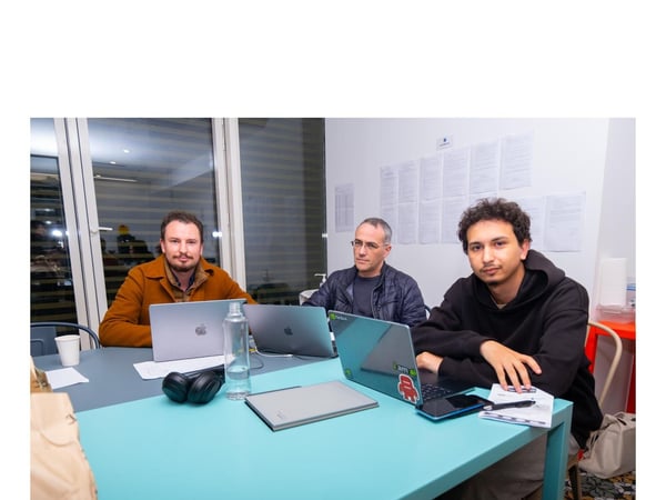 Three men are seated closely at a bright teal and gray table, actively working on open laptops in an indoor setting, likely an office or collaborative workspace.