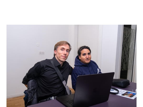 Two men are seated side-by-side at a purple table indoors, looking directly at the camera, with a black laptop positioned in front of them.