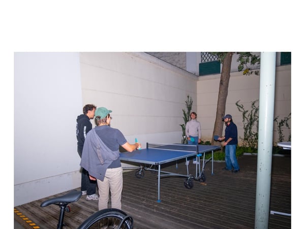 A photograph capturing four people gathered around a blue table tennis table in an outdoor courtyard or patio with wooden decking.