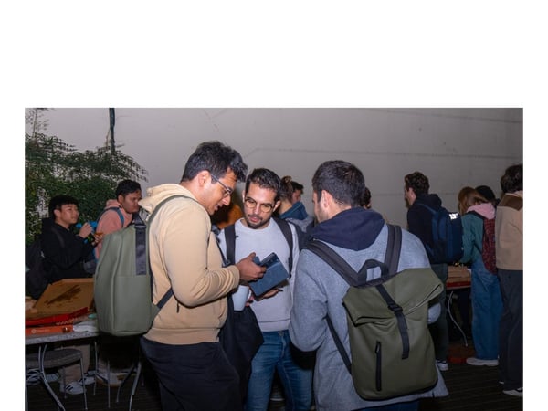 A candid photograph captures a group of young people socializing outdoors during the evening, with three men in the foreground focused on looking at a small electronic device.
