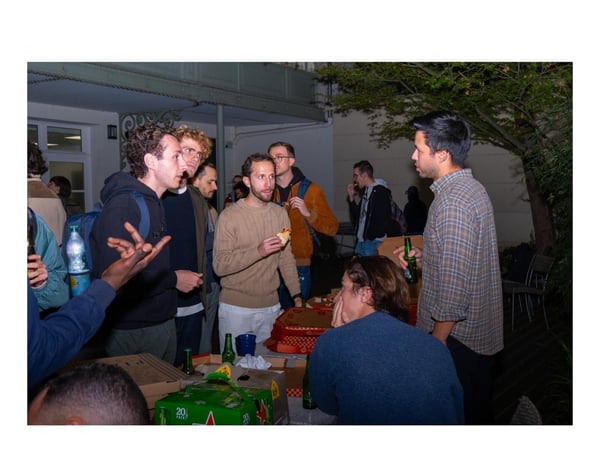 A candid photograph taken at night showing a group of young men socializing outdoors in a courtyard, gathered around a table with pizza and a case of beer.