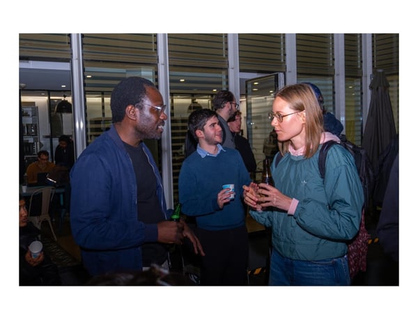 A candid photograph taken indoors at night shows a group of people socializing, focusing on three individuals engaged in conversation while holding drinks.