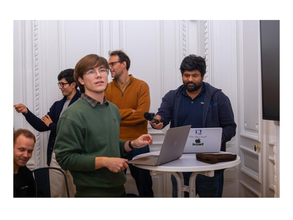 A candid photograph showing five men gathered in a formal room, likely during a technical presentation or discussion, with two men standing near a small table holding open laptops and a microphone.