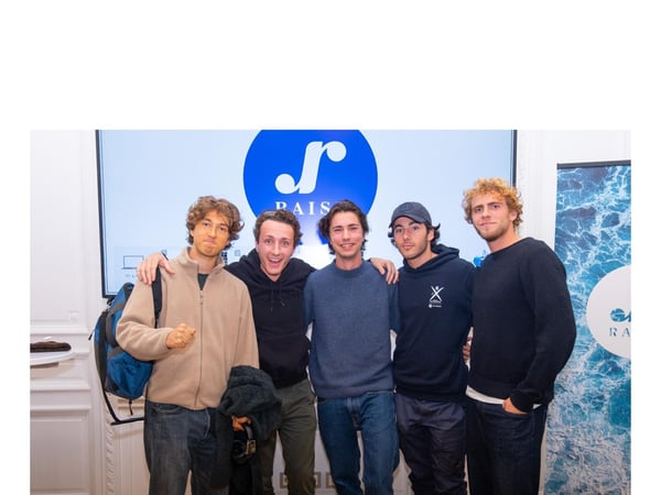 Five young men are posing closely together for a group photo indoors, standing in front of a large screen displaying a blue circular logo and the partial text "PAIS."