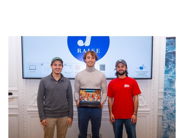 A photograph of three young men standing side-by-side in a formal indoor setting, posing in front of a large screen displaying the 'RAISE' logo and technical connection instructions.