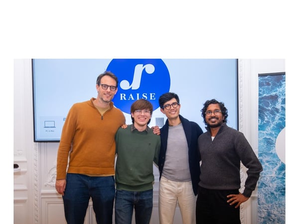 Four men are standing together, posing for a group photograph indoors in front of a large screen displaying the logo and name "RAISE."