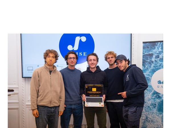 Five young men are posing for a group photo indoors, standing in front of a large screen displaying a blue circular logo and the text 'ISE,' while one of them holds an open laptop showing a project title.