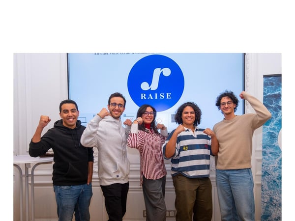 A group photograph of five young people standing side-by-side indoors, posing enthusiastically with raised fists in front of a large screen displaying the blue and white 'RAISE' logo.
