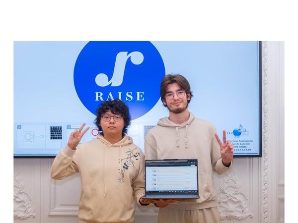 A photograph of two young men standing side-by-side in front of a large screen displaying the 'RAISE' logo and holding a laptop, both making a peace sign gesture.