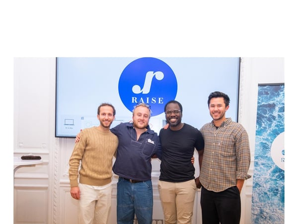 Four men stand side-by-side, smiling for a group photo in a brightly lit indoor setting, with a large screen displaying a blue circular logo and the word "RAISE" behind them.