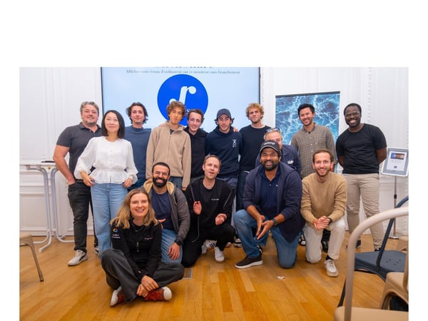 A group of 15 colleagues, likely a startup team, poses for a group photo indoors, arranged in standing and kneeling rows in front of a screen displaying a blue logo and French text.