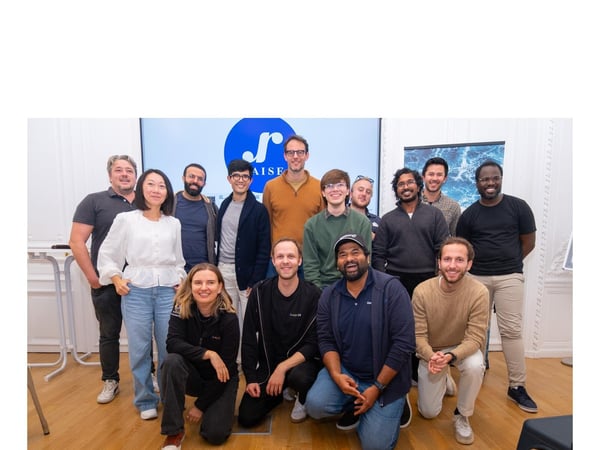 A group of thirteen people, likely colleagues or a team, pose for a group photo indoors in a bright room with light wood flooring, standing and kneeling in front of a large screen displaying a blue and white logo and the text "AISE[partial]".
