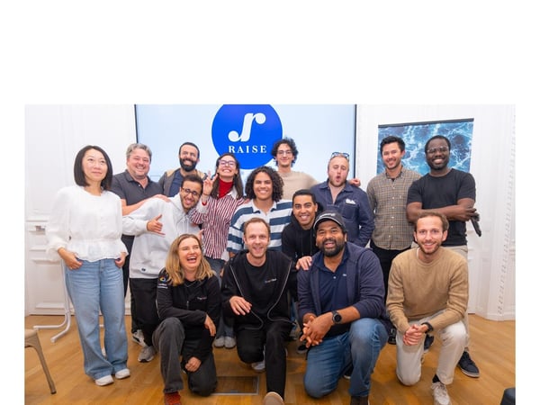 A group portrait of fifteen people, likely a team or cohort, posing indoors in front of a large screen displaying the blue and white "RAISE" logo.