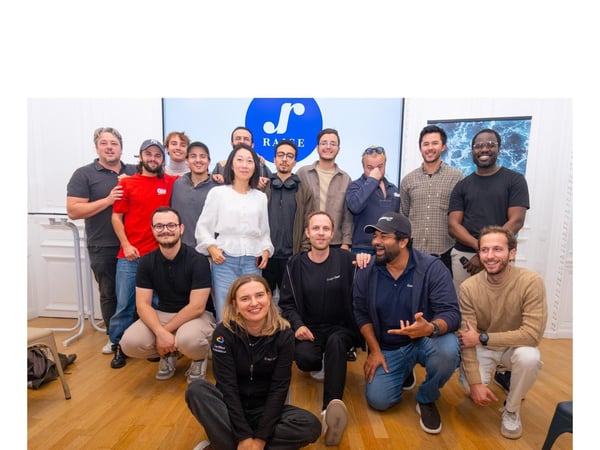 A group of about fifteen young professionals, composed primarily of men, poses for a team photo indoors, standing and kneeling in front of a large screen displaying a blue and white logo with the text "RACE."