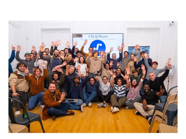 A large group of approximately 38 people, likely a corporate team, poses for a group photo indoors, smiling and enthusiastically waving their hands in the air. They are standing and sitting in front of a screen displaying the company name "ClickShare" and its logo.