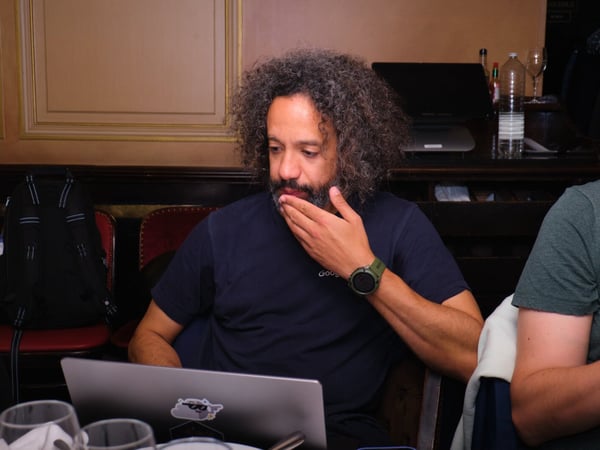 A man with voluminous curly hair and a beard sits at a table in an ornate, dimly lit room, intently looking down at his open laptop.