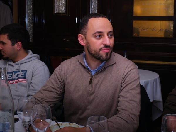 A candid photograph of three men seated around a dining table in a dark, formal indoor setting, likely a restaurant, with the central subject wearing a brown quarter-zip sweater.