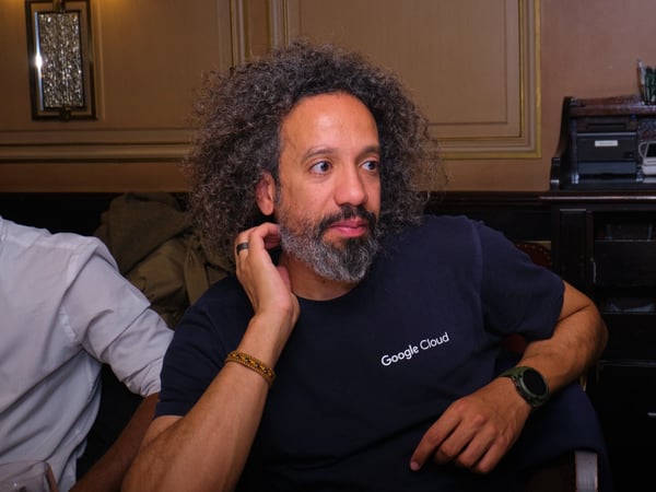 A close-up photograph of a man with large, curly graying hair and a beard, wearing a dark t-shirt featuring the text "Google Cloud." He is sitting indoors in a dimly lit, warm-toned setting, looking off to the side while resting his hand near his chin.