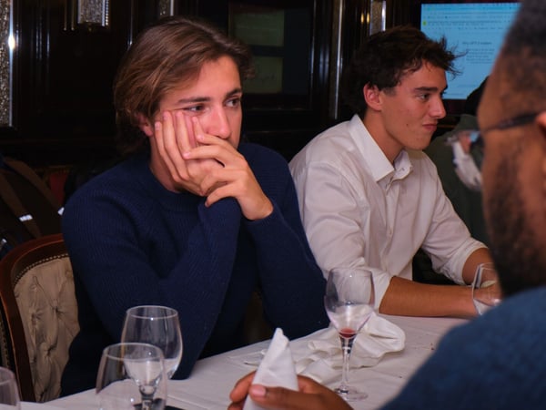 A candid indoor photograph capturing three young men seated around a table, likely during a dinner or meeting, with the central figure resting his face contemplatively on his hands.