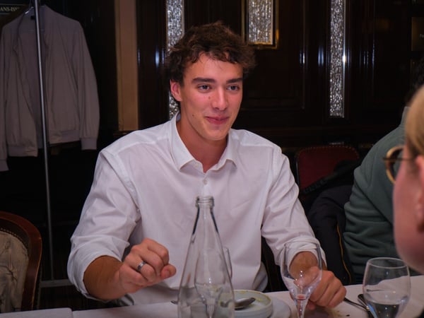 A young man wearing a white collared shirt sits at a dining table in a dimly lit, traditional restaurant, smiling and gesturing during a conversation.