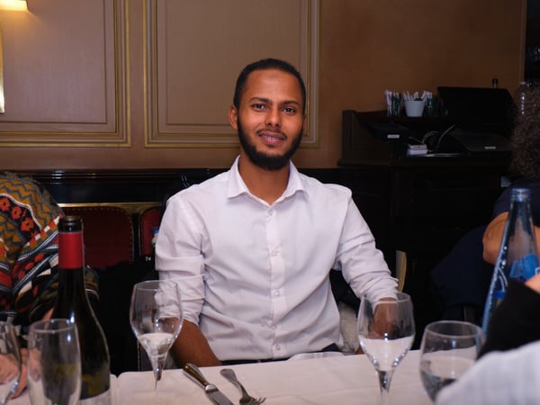 A portrait-style photograph of a smiling man wearing a white collared shirt, seated at a formal dining table in a dimly lit, elegant restaurant setting.
