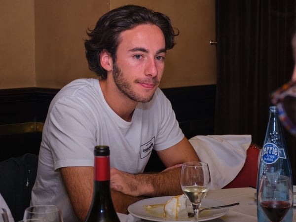 A candid photograph captures a young man with dark hair and a beard, wearing a white t-shirt, sitting at a table in a dimly lit indoor restaurant setting. The table is set with wine glasses, a bottle of Perrier, a bottle of red wine, and a plate containing dessert.