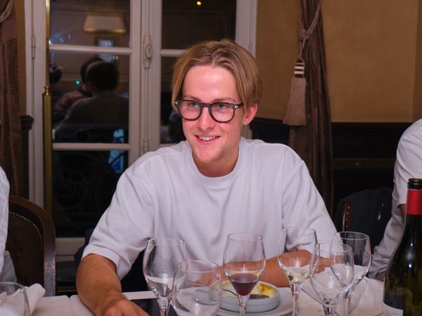 A candid indoor photo showing a young man wearing glasses and a white t-shirt, smiling while seated at a formal dinner table with multiple wine glasses and a wine bottle.