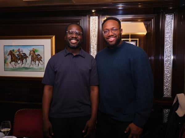 Two Black men wearing glasses stand side-by-side and smile directly at the camera in a dimly lit, upscale indoor setting featuring dark wood paneling and a framed painting of a horse race.