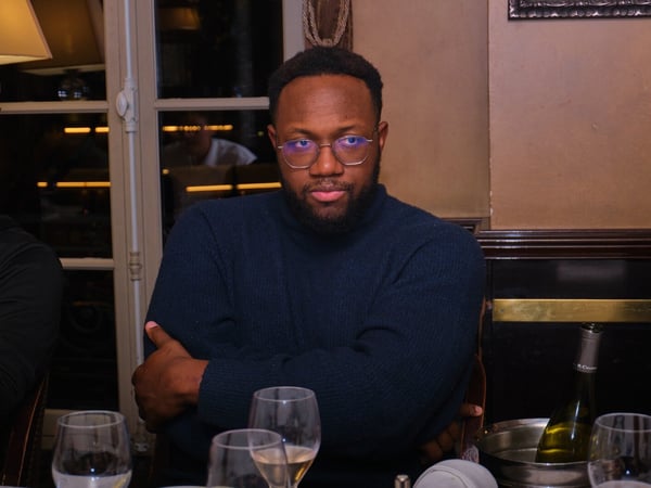 A close-up photograph of a man wearing glasses and a dark blue turtleneck sweater, seated at a dining table with his arms crossed in a dimly lit, warm-toned restaurant setting.