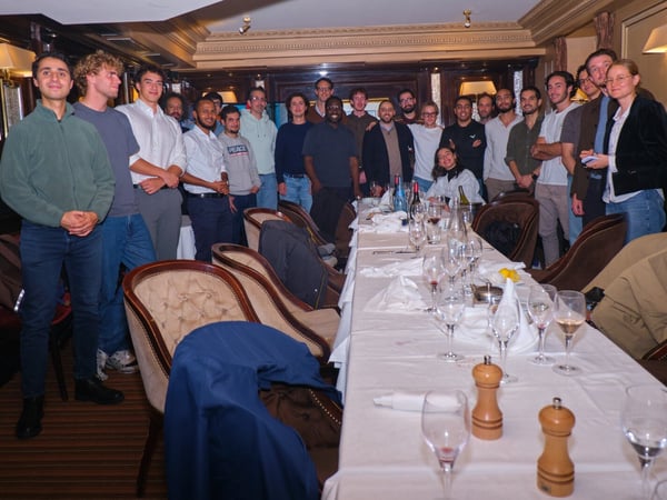 A large group of approximately twenty young adults, predominantly men, poses for a group photo behind a long dining table set with wine glasses and bottles in a formal, wood-paneled restaurant setting.
