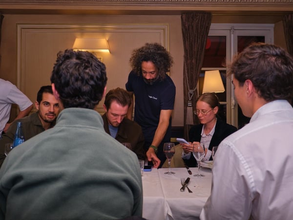 A candid photograph captures a group of people gathered around a white-clothed dining table in a dimly lit, formal indoor setting, possibly discussing something on a mobile phone.