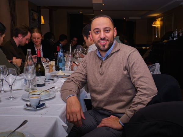 A man wearing a beige quarter-zip sweater and a beard is seated at a white-clothed dining table in a dimly lit restaurant, smiling directly at the camera. Several other people are visible seated around the table in the background, which is cluttered with wine bottles, glasses, and used dishes.