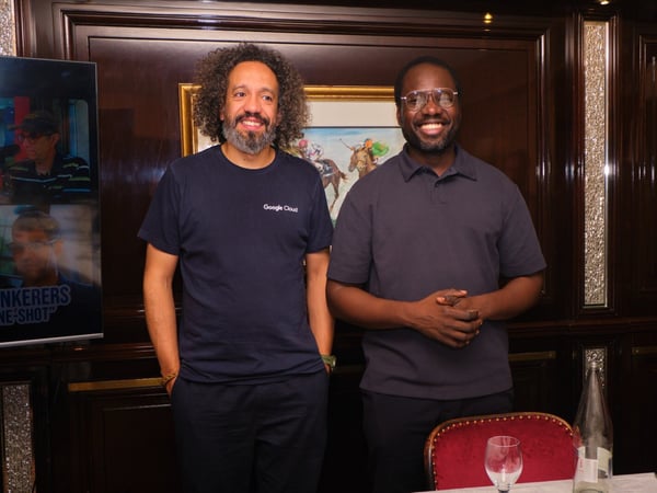Two men are standing side-by-side and smiling at the camera in what appears to be a formal indoor setting with dark wood paneling and elegant decor.