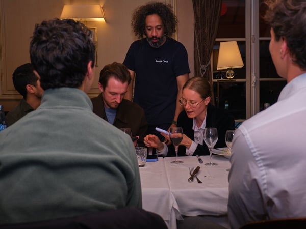 A group of five people is gathered around a white-clothed dining table in a dimly lit, formal indoor setting, possibly a restaurant, with several individuals looking at their mobile phones. One man is standing behind the table wearing a dark t-shirt with the 'Google Cloud' logo.