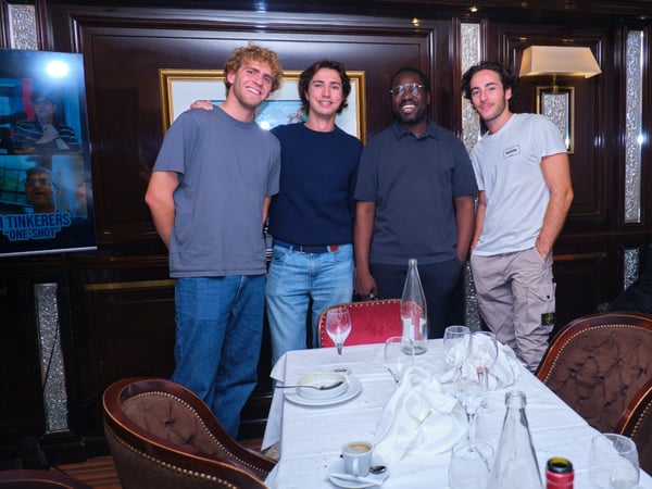 Four men are standing together, smiling, behind a set dining table in a dimly lit, wood-paneled restaurant or private dining room. A poster or screen advertising the title "TINKERERS ONE-SHOT" is visible on the left side of the frame.