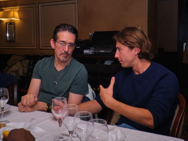 A photograph showing two men seated at a dining table in a dimly lit restaurant, engaged in a conversation, with the younger man on the right gesturing with his hand.