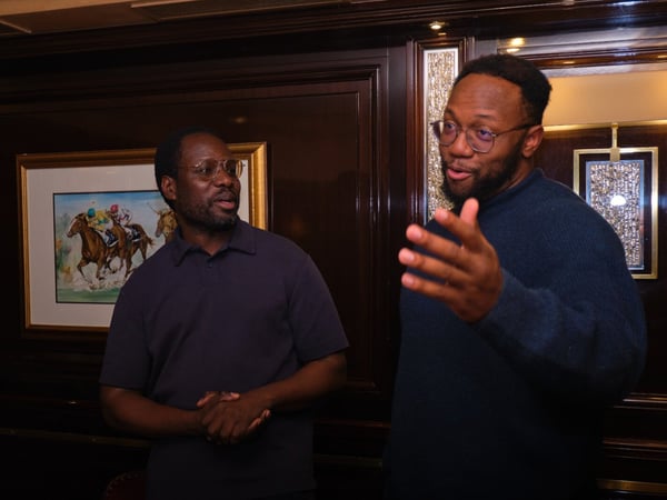 Two men, both wearing glasses, are standing in a dimly lit, upscale room with dark wood paneling and framed artwork, engaged in an animated discussion.