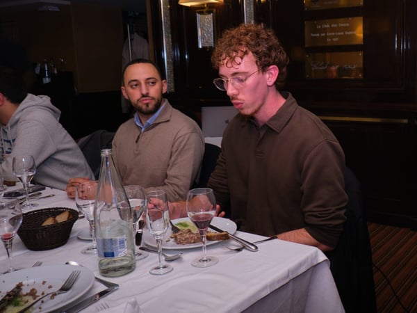A candid indoor photograph capturing several people seated around a formal dining table, focusing on two men engaged in conversation or contemplation during a meal.
