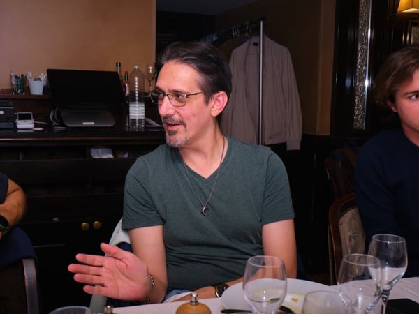 A candid photograph of a man wearing glasses and a green shirt, seated at a dining table in a dimly lit restaurant, gesturing with his hand while participating in a conversation.