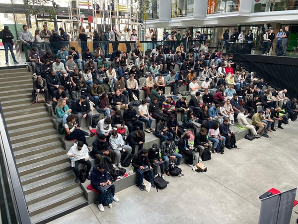 A wide-angle photograph captures a large audience of students or attendees seated closely together on tiered concrete steps inside a modern, multi-story building atrium, likely attending a presentation or lecture.