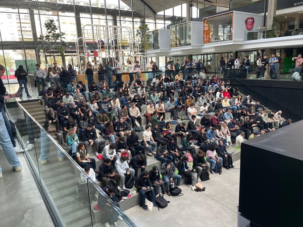 A large crowd of people is seated in a modern, tiered amphitheater-style seating area within a spacious, glass-walled atrium, likely attending a lecture or event.