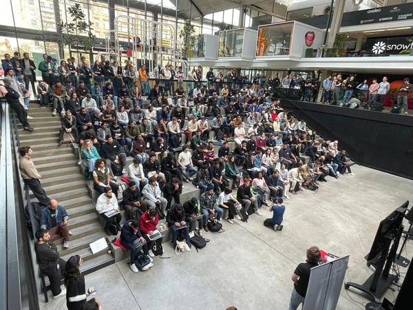 A high-angle photograph captures a large audience seated on tiered concrete steps inside a modern, brightly lit atrium or auditorium, attending a presentation or event.