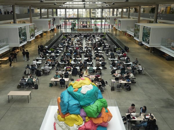 A wide-angle photograph captures a massive indoor atrium, likely during a hackathon or large co-working event, where hundreds of people are seated at rows of tables working on laptops, with a large, colorful, piled-up sculpture dominating the foreground.