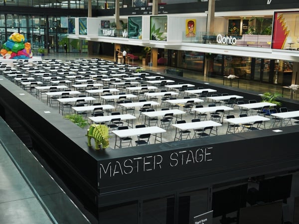 A high-angle photograph capturing the interior of a large, modern, multi-story building atrium set up for a massive event, featuring hundreds of empty white tables and black chairs arranged on a raised platform labeled "MASTER STAGE."