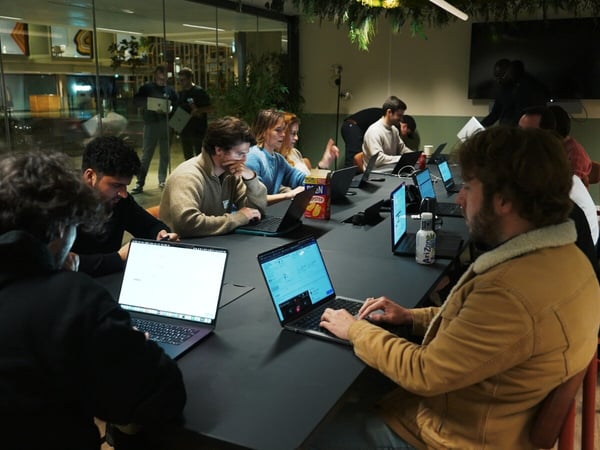 A group of people are seated around a large, dark table working on laptops in a dimly lit modern office space, suggesting a late-day work session or coding event.