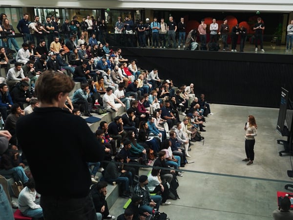 A high-angle photograph captures a large indoor event where a female presenter addresses a sizable audience seated on tiered steps in a modern, multi-level venue. The upper level features a black railing adorned with the text 'MONSTER STAGE' and a neon green wireframe dinosaur sculpture.