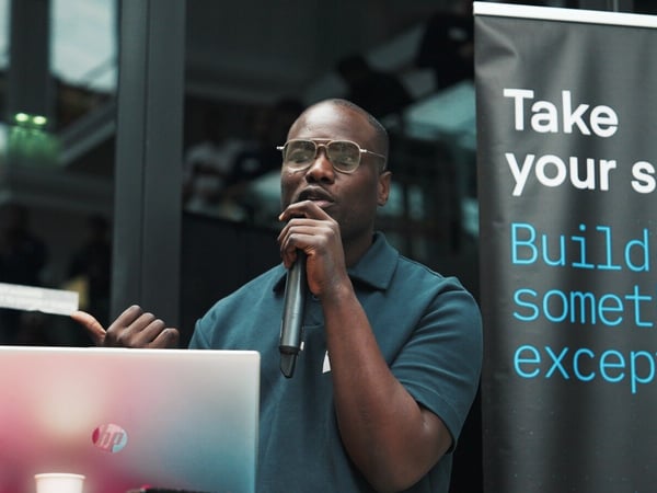 A dark, candid photograph captures a man wearing glasses and a polo shirt speaking into a microphone during an indoor presentation, standing behind a laptop with a promotional banner visible on the right.